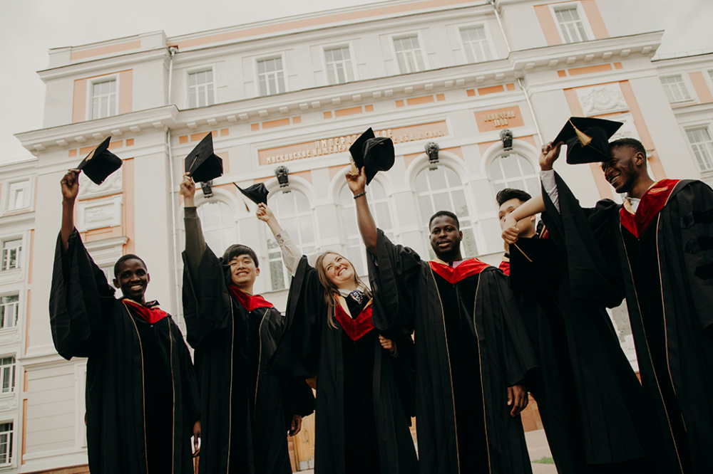 A group of six graduates wearing black robes and red stoles hold their caps in the air in front of a grand building.